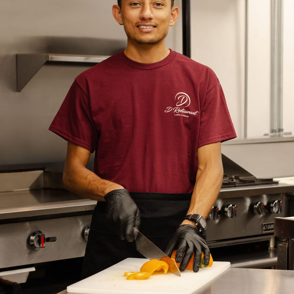 Restaurant worker in a maroon shirt slices yellow bell pepper on a cutting board in a commercial kitchen.
