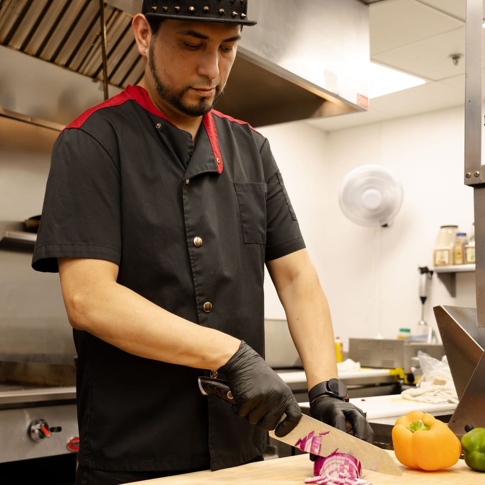 Chef in a commercial kitchen slices red onion on a wooden cutting board beside orange and green bell peppers.