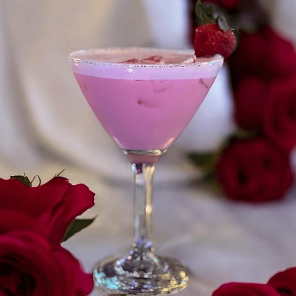 Pink cocktail in a martini glass, garnished with a strawberry, surrounded by red roses.