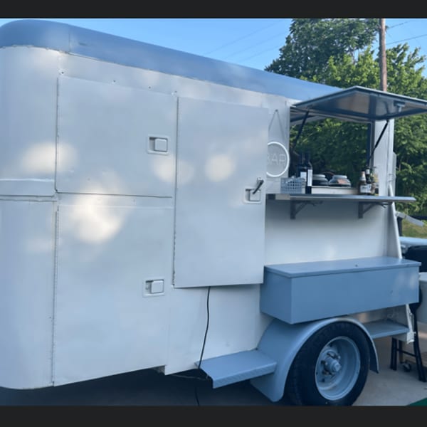 A white and gray mobile bar trailer with an open serving window, bottles, and a neon "BAR" sign.