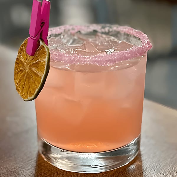 Pink cocktail with a sugar-rimmed glass, garnished with a dried orange slice and a pink clothespin.