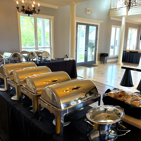 A buffet line with several chafing dishes and a basket of bread rolls in a bright room.