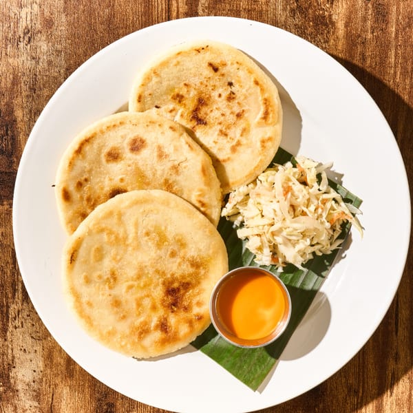 Three golden-brown pupusas with curtido and a small cup of orange sauce on a white plate.
