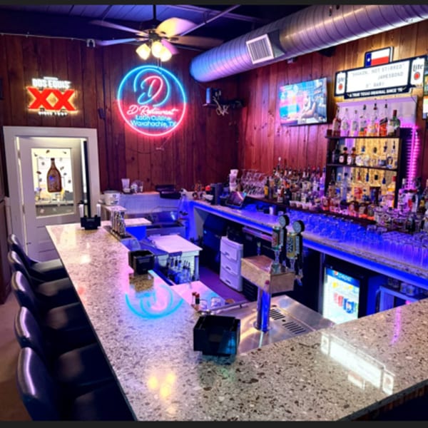 A vibrant bar with neon signs, a fully stocked liquor shelf, and blue under-counter lighting.