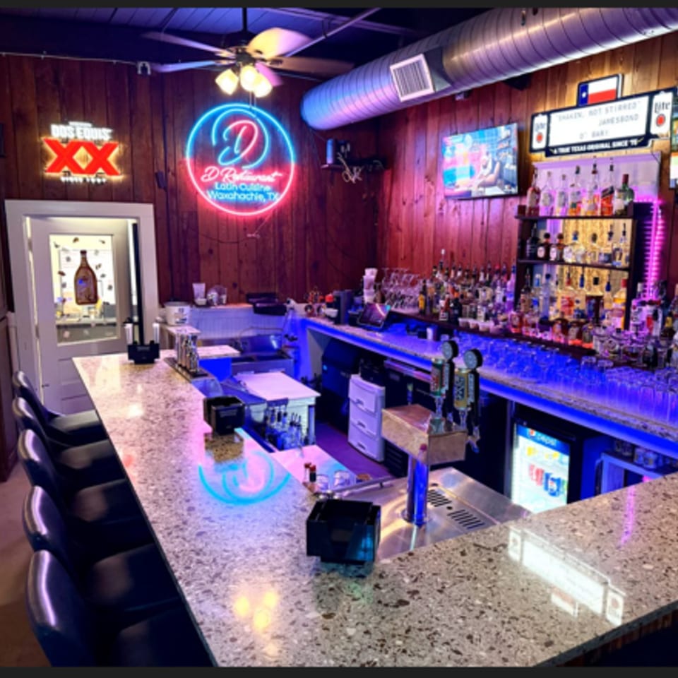 A vibrant bar with neon signs, a fully stocked liquor shelf, and blue under-counter lighting.