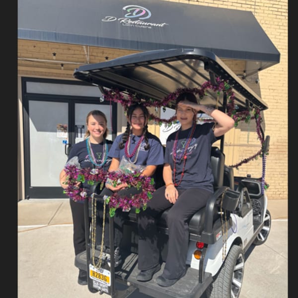 Three young women in D Restaurant shirts and Mardi Gras beads pose in a decorated golf cart.