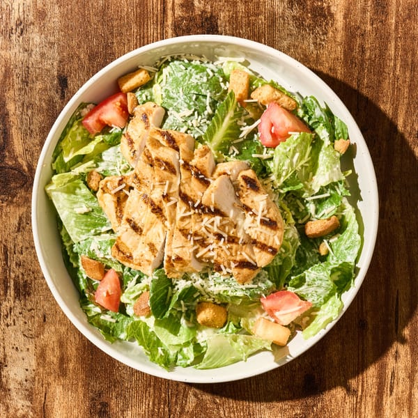 Grilled chicken Caesar salad with romaine, croutons, tomatoes, and parmesan cheese in a white bowl.