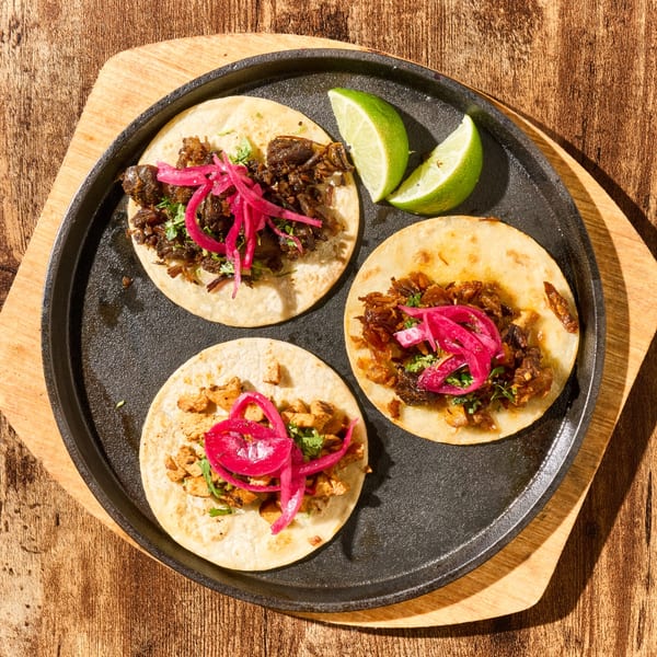 Three Latin-American tacos with meat, cilantro, and pickled red onions, served with lime wedges.